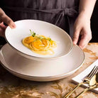 Gold-rimmed flat plate in white, lifestyle shot showing the plate with pasta on a styled table