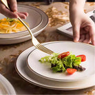 Gold-rimmed flat plate in white, lifestyle shot showing a plated meal being served at a table